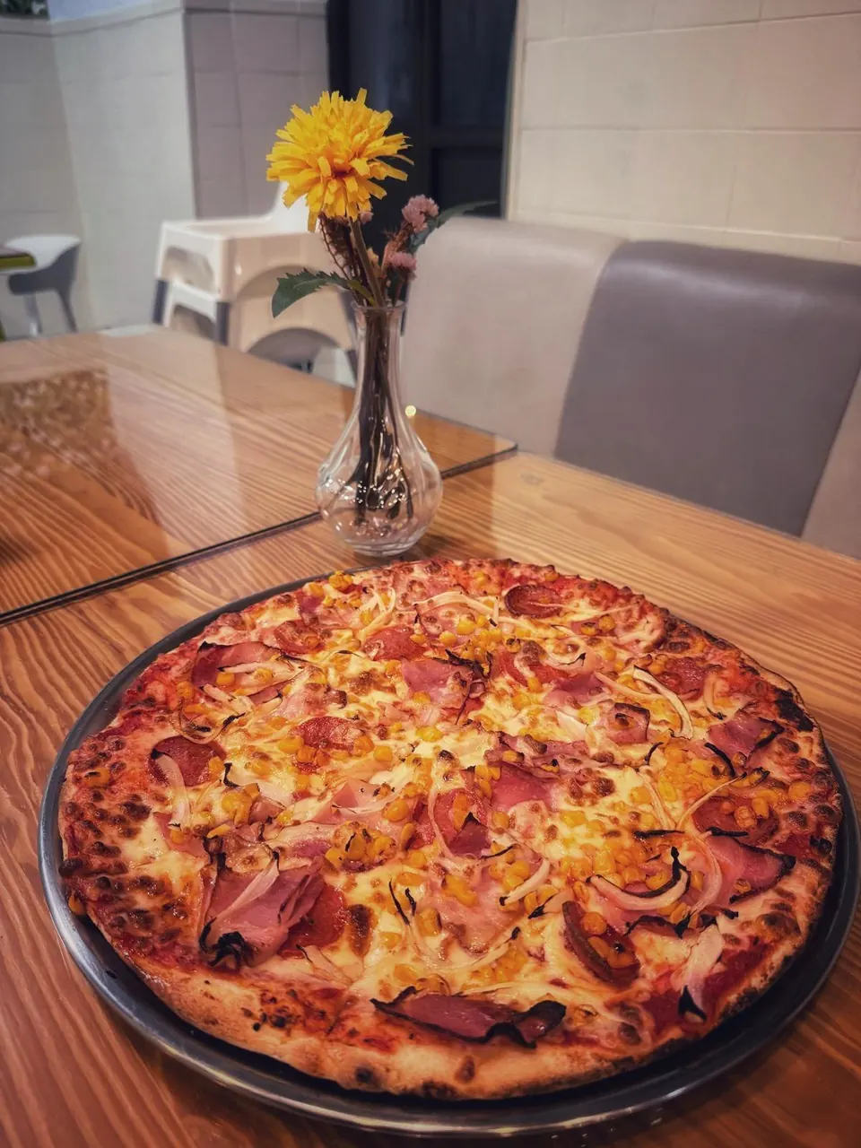 Pizza de jamón, maíz y cebolla servida en la mesa con un jarrón de flores detrás