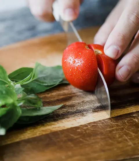 Manos cortando un tomate sobre una tabla de madera, junto a unas hojas de albahaca